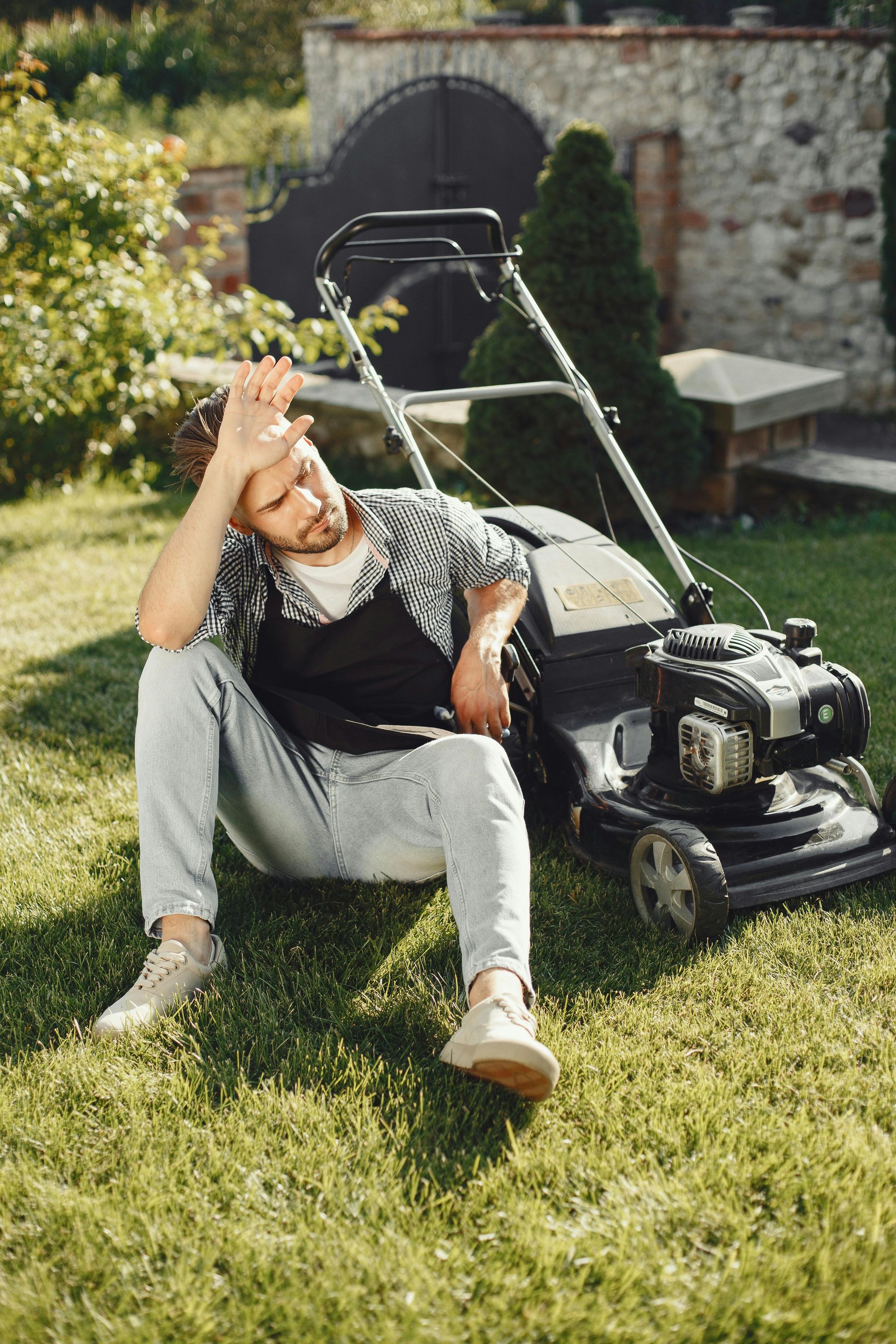 A tired man sitting on the grass with a lawn mower after gardening.